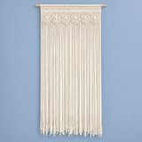 An off-white macrame wall hanging with intricate woven patterns at the top and long hanging fringe below, suspended from a light natural wood rod against a solid light blue wall.