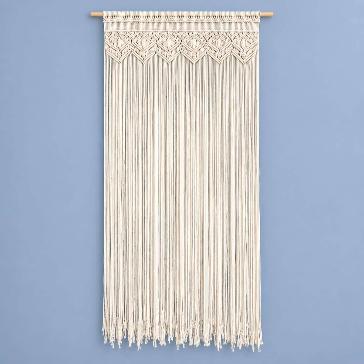 An off-white macrame wall hanging with intricate woven patterns at the top and long hanging fringe below, suspended from a light natural wood rod against a solid light blue wall.