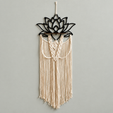 A bohemian-style macrame wall hanging featuring a black metal lotus flower frame at the top and cream-colored woven cord fringe below, hung against a plain light gray wall.