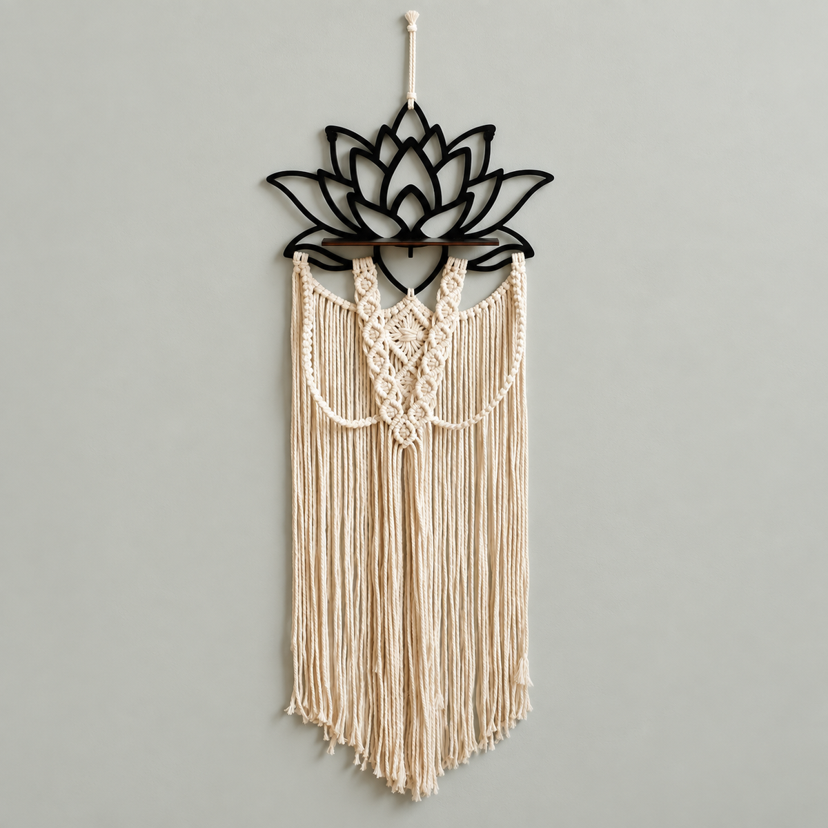 A bohemian-style macrame wall hanging featuring a black metal lotus flower frame at the top and cream-colored woven cord fringe below, hung against a plain light gray wall.