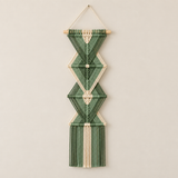 A vertical macrame wall hanging with a geometric diamond pattern, made from green and cream cotton cord on a light wood dowel, hanging against a plain beige wall.