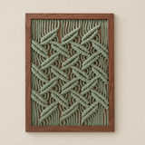 A framed wall art piece featuring a textured, woven geometric pattern in sage green rope or cord over a medium brown wooden frame, displayed against a warm beige wall.