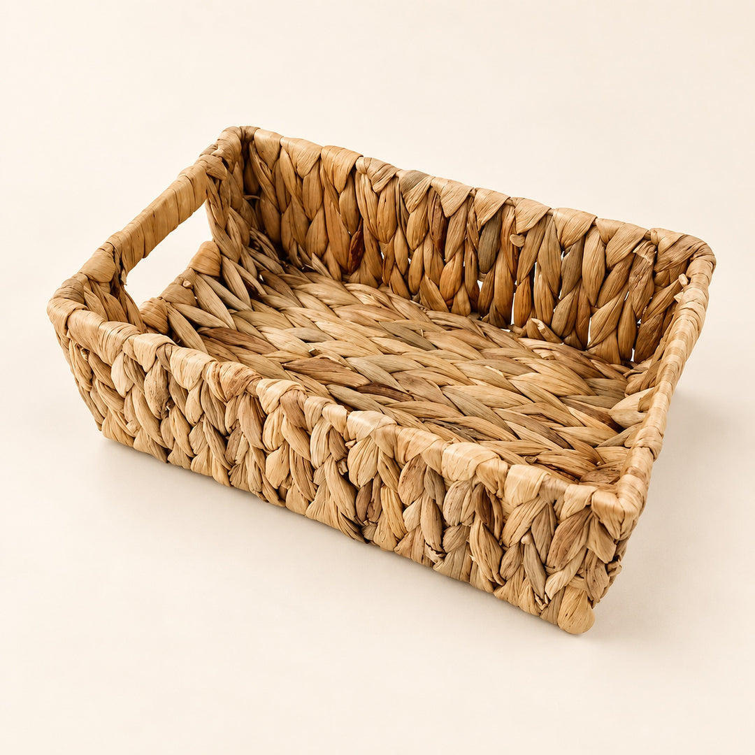 A rectangular woven seagrass or straw basket tray with integrated side handles, shown on a clean, solid, warm off-white background.
