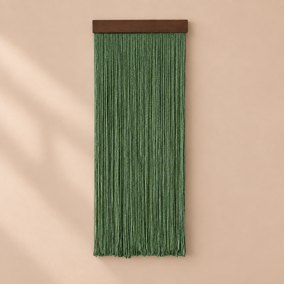 A green fringe wall hanging with a dark brown wooden top bar, displayed against a warm beige wall with soft diagonal shadows.