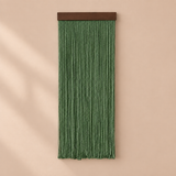 A green fringe wall hanging with a dark brown wooden top bar, displayed against a warm beige wall with soft diagonal shadows.
