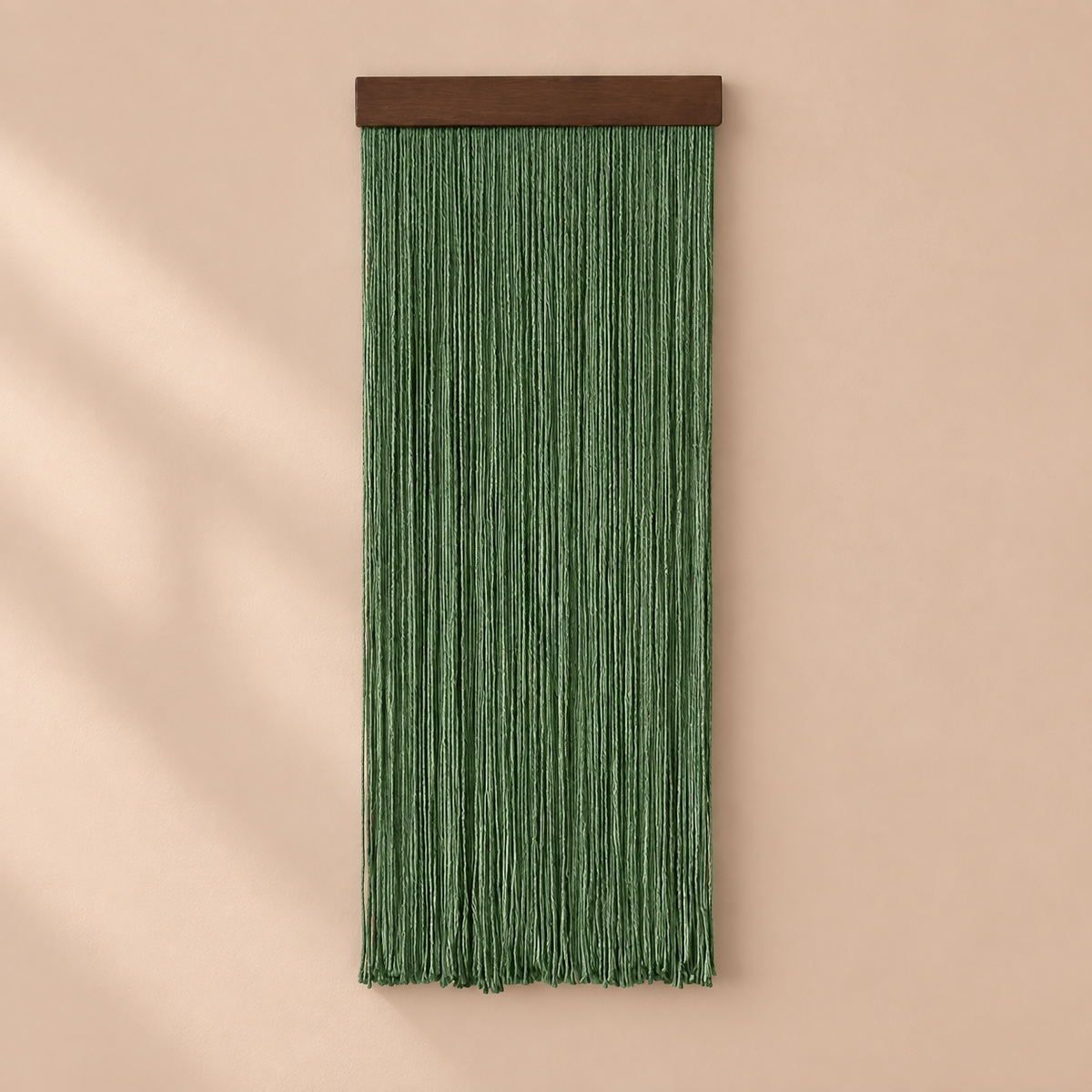 A green fringe wall hanging with a dark brown wooden top bar, displayed against a warm beige wall with soft diagonal shadows.
