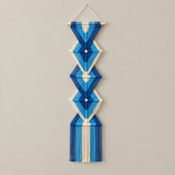 A vertical macrame wall hanging with geometric diamond and triangle patterns, made from blue and white threads on a light wooden dowel, hanging against a plain light gray wall.