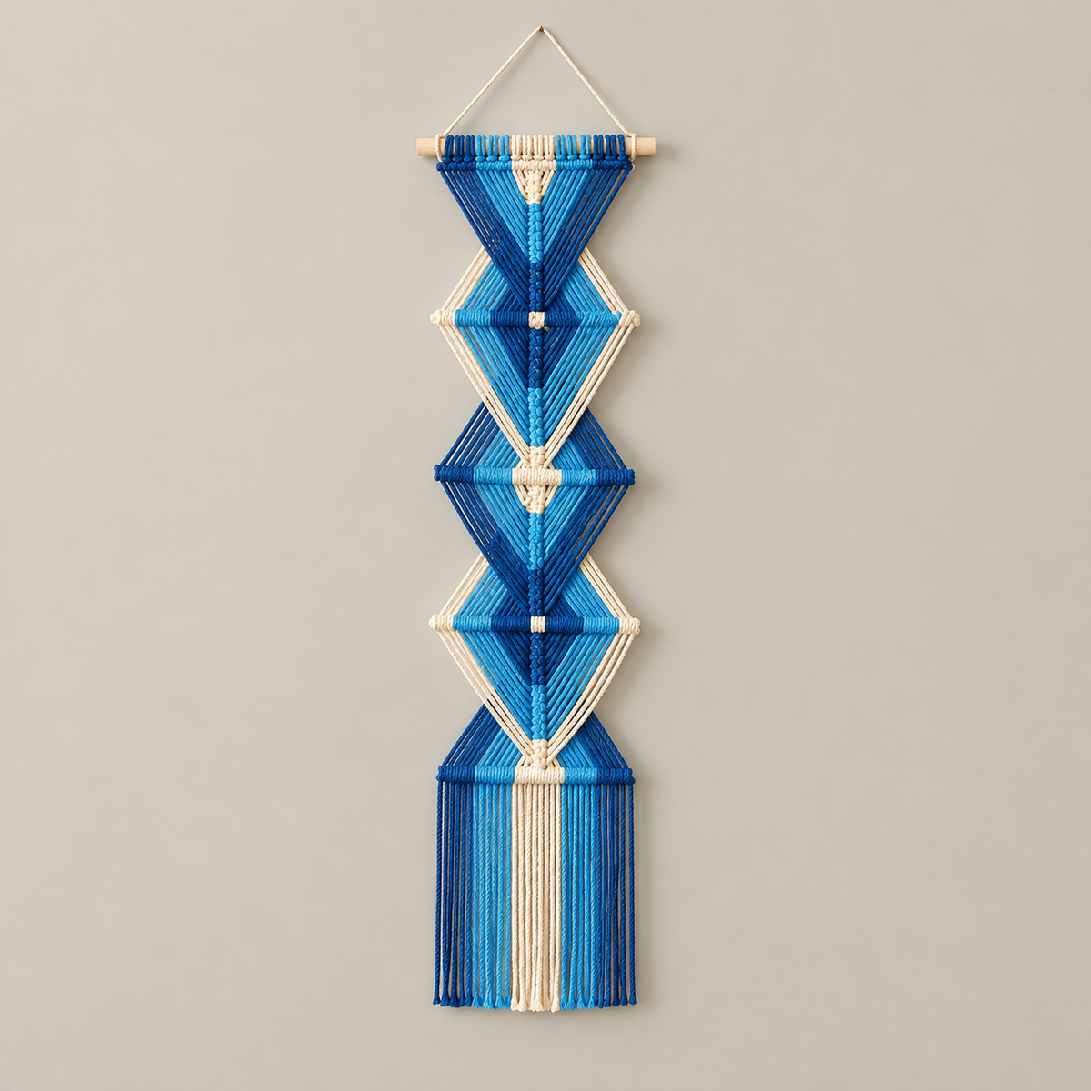 A vertical macrame wall hanging with geometric diamond and triangle patterns, made from blue and white threads on a light wooden dowel, hanging against a plain light gray wall.