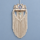 A bohemian-style wall hanging featuring a natural wood lotus-shaped top, cream macrame fringe, and two small hanging crystals, one pink and one light green, set against a solid blue background.