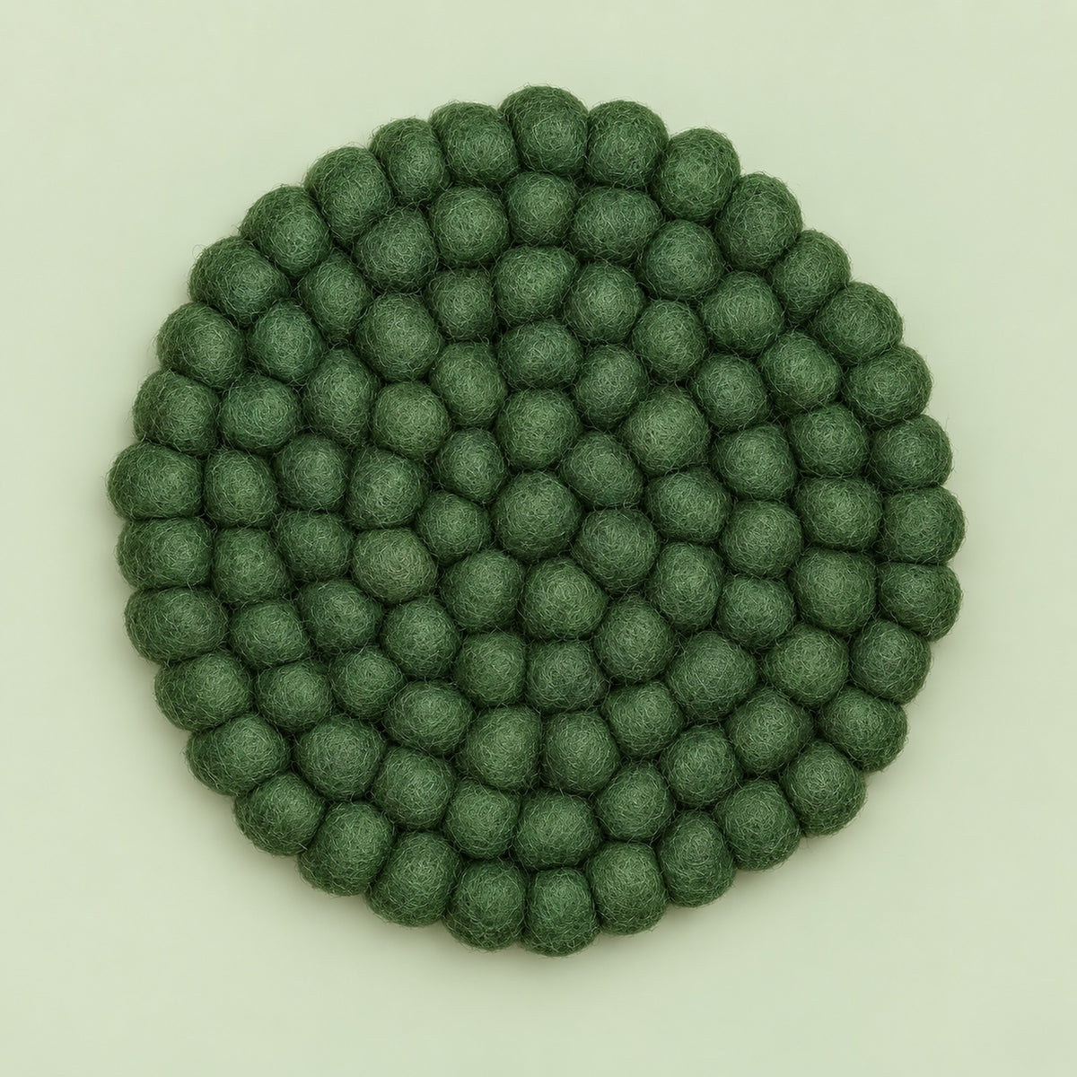 A round felt mat made of small textured pom-pom balls in a muted sage green color, photographed from directly above on a light off-white background.