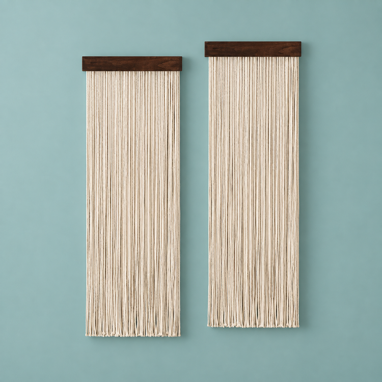 Two vertical wall hanging pieces with dark wood tops and long cream string fringe, displayed against a muted blue-green wall.