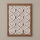 A square framed macrame wall art piece featuring an intricate cream-colored woven geometric pattern inside a medium brown wooden frame, mounted on a neutral beige wall.
