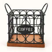 A black metal wire coffee basket with decorative oval wire patterns, a wooden base, and two side handles. A black oval sign on the front reads 'COFFEE' in white letters, set against a plain white background.