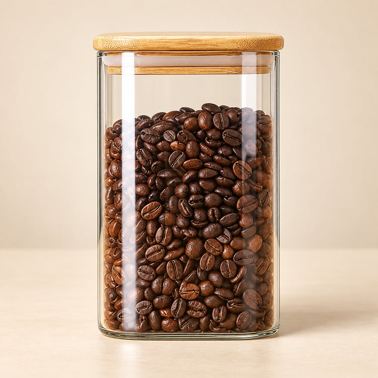 A clear glass storage jar filled with roasted coffee beans, topped with a light bamboo lid, set against a warm beige background.