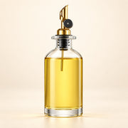 A clear glass bottle filled with golden yellow oil, featuring a polished gold metal pour spout with a black collar, set against a warm beige background.