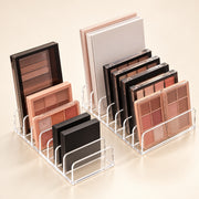 Two clear acrylic makeup organizer stands holding multiple eyeshadow palettes and compact powders in neutral and pink tones, arranged on a warm beige studio background.