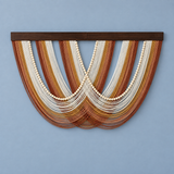 A bohemian-style wall hanging featuring multiple curved strands of brown, tan, and cream cord with central rows of white wooden beads, suspended from a dark wood top bar against a light blue wall.