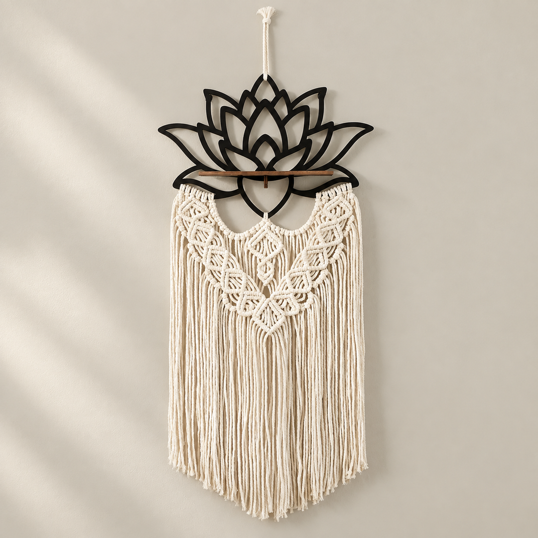 A bohemian-style macrame wall hanging with a black metal lotus flower frame at the top, a wooden dowel, and intricate cream-colored woven fringes, hanging on a neutral beige wall.