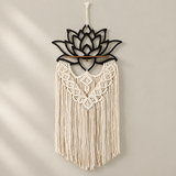 A bohemian-style macrame wall hanging with a black metal lotus flower frame at the top, a wooden dowel, and intricate cream-colored woven fringes, hanging on a neutral beige wall.