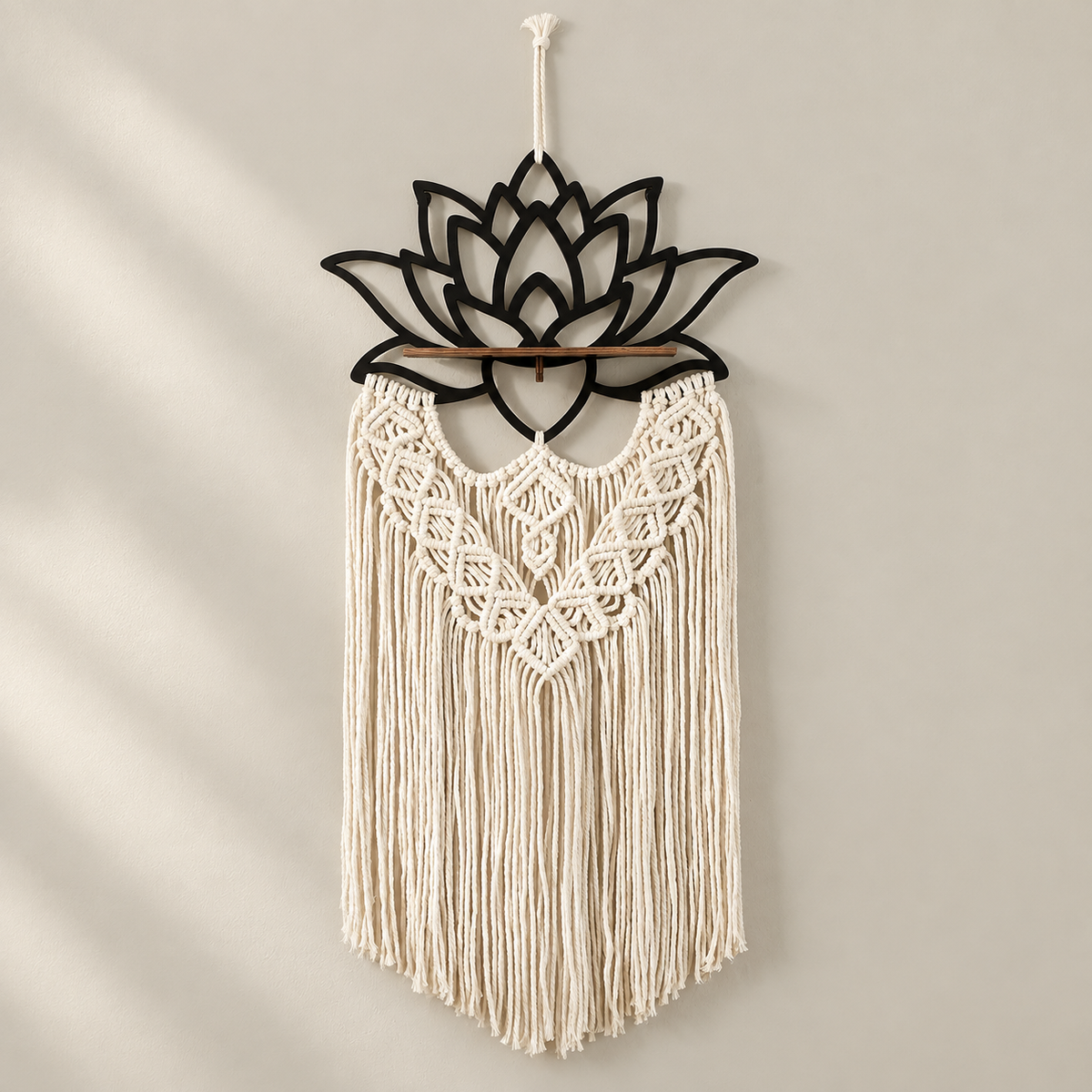 A bohemian-style macrame wall hanging with a black metal lotus flower frame at the top, a wooden dowel, and intricate cream-colored woven fringes, hanging on a neutral beige wall.
