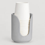 A light gray ribbed cup holder or cup dispenser holding a stack of white paper cups, photographed on a clean light gray background.