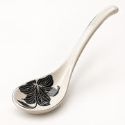 An off-white ceramic serving spoon featuring a black floral pattern on the bowl, set against a clean white background.