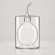 Clear glass decanter with a rounded bottle body, rectangular outer container, and a curved pouring spout, photographed on a light gray studio background.