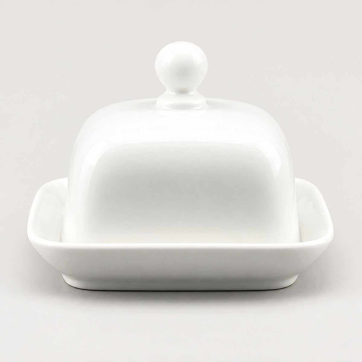 A white ceramic butter dish with a domed lid featuring a round knob and a shallow square base, photographed on a clean light gray background.