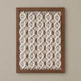 A framed macrame wall hanging with an intricate cream-colored woven geometric pattern inside a medium brown wooden frame, displayed against a solid beige wall.