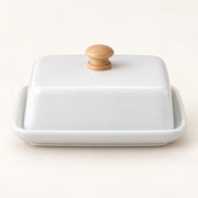 A white ceramic butter dish with a matching lid and a round light wood knob, set against a plain off-white background.