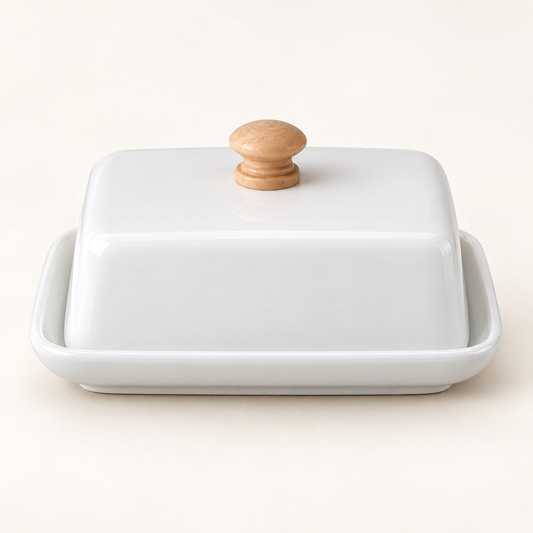 A white ceramic butter dish with a matching lid and a round light wood knob, set against a plain off-white background.