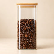 A tall, clear glass coffee canister with a light natural wood lid, filled with roasted brown coffee beans, set against a warm beige background.