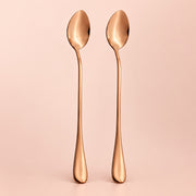 Two long-handled spoons with a polished rose gold metallic finish, standing upright side-by-side against a solid pale pink background.