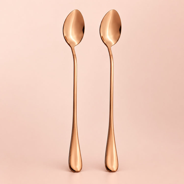 Two long-handled spoons with a polished rose gold metallic finish, standing upright side-by-side against a solid pale pink background.