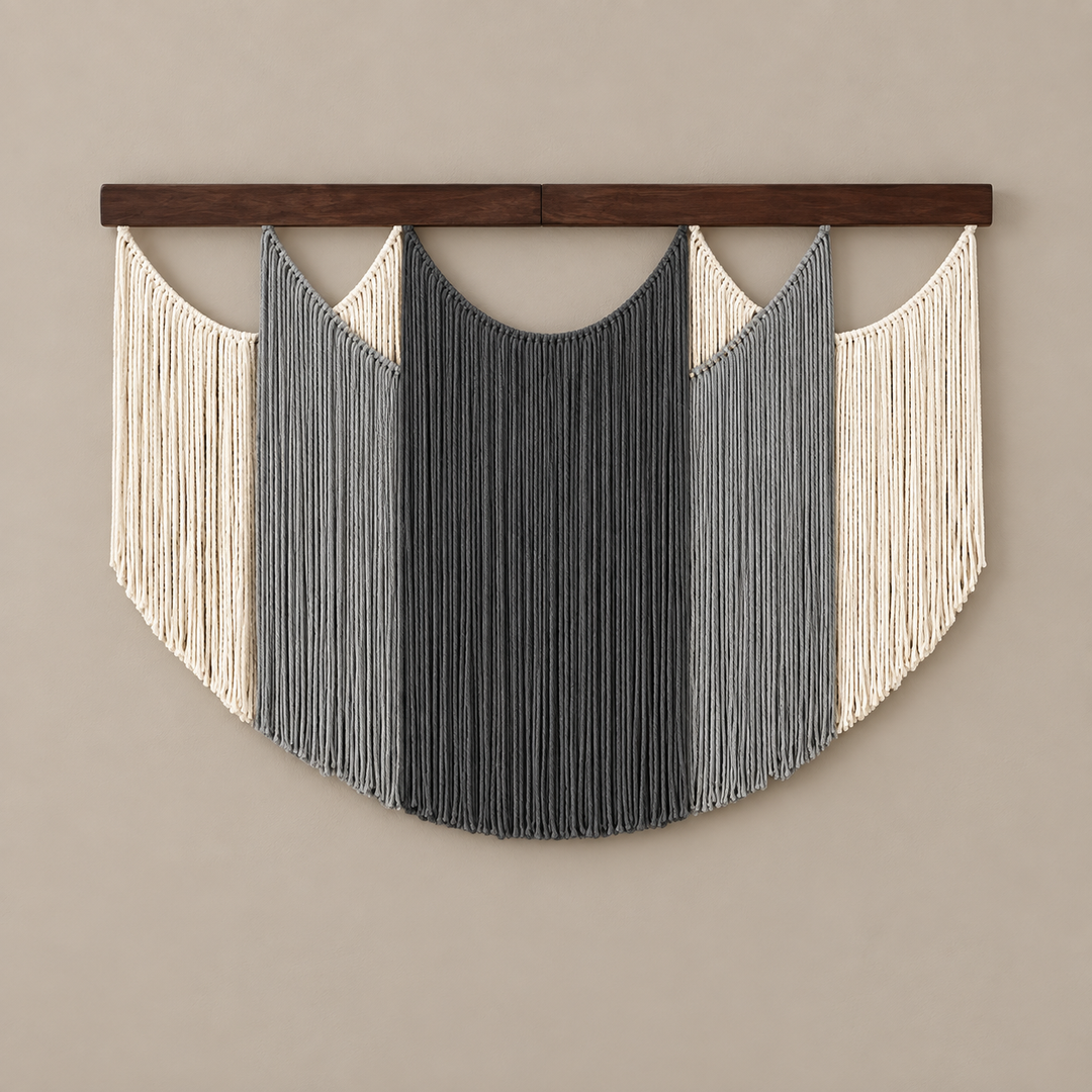 A bohemian-style macrame wall hanging with a dark wood top bar, featuring vertical fringe in cream, light gray, and charcoal gray, suspended against a solid beige wall.