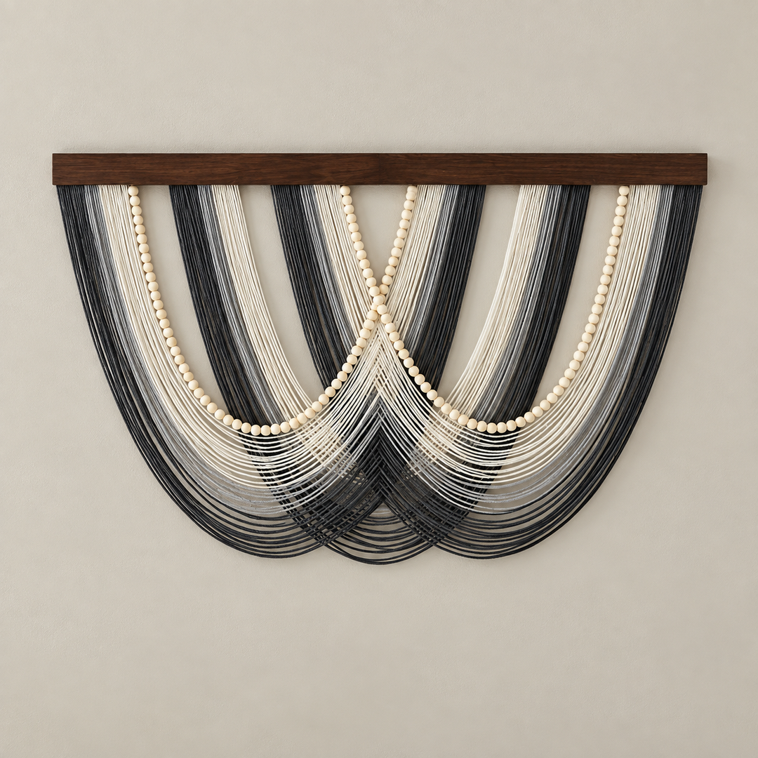 A wall-mounted decorative textile or wall hanging with a dark wooden top bar, featuring layered strands in black, gray, white, and cream arranged in a fan shape against a plain beige wall.