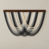 A wall-mounted decorative textile or wall hanging with a dark wooden top bar, featuring layered strands in black, gray, white, and cream arranged in a fan shape against a plain beige wall.