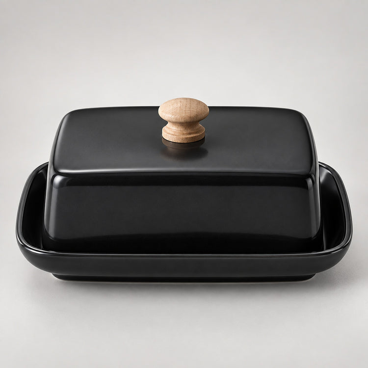 A glossy black ceramic butter dish with a matching lid and a round light wood knob, set against a neutral light gray background.