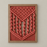 A framed macrame wall hanging with a geometric pattern in a coral pink cord, set within a light brown wooden frame against a neutral beige wall.