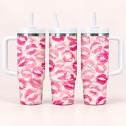 Three handled stainless steel tumblers with white lids and straws, decorated with a repeating pink lip print pattern on a light cream background, shown against a pale pink studio backdrop.