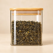 A clear glass jar with a light natural wood lid, filled with rolled green tea leaves, set against a warm beige background.