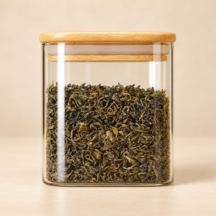 A clear glass jar with a light natural wood lid, filled with rolled green tea leaves, set against a warm beige background.