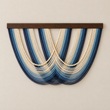 A wall-mounted decorative textile wall hanging with multiple draped strands in shades of blue and cream, overlaid with three vertical rows of cream beads, suspended from a dark wood bar against a light beige wall.