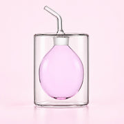 A clear glass double-walled tumbler with a matching clear glass pour spout lid and angled straw, filled with light pink liquid, set against a solid pale pink background.