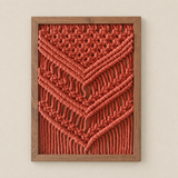 A framed macrame wall art piece featuring a textured geometric chevron pattern in terracotta rope, enclosed in a natural wood frame and mounted on a light beige wall.