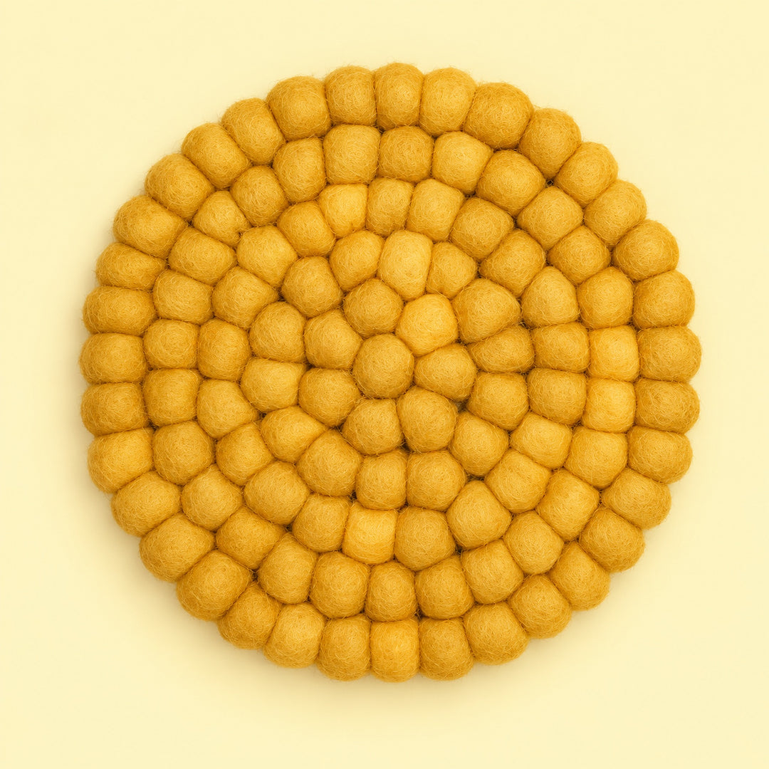 A round felt placemat or coaster made of small textured pom-pom balls, arranged in concentric circles, in a warm mustard yellow color on a light cream background.