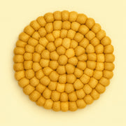 A round felt placemat or coaster made of small textured pom-pom balls, arranged in concentric circles, in a warm mustard yellow color on a light cream background.