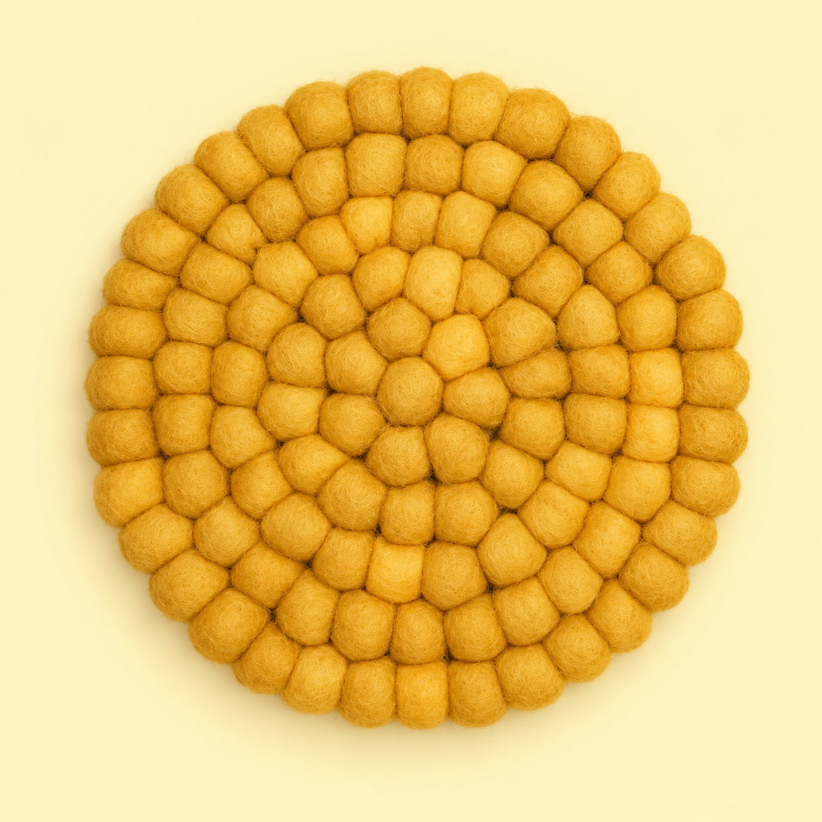 A round felt placemat or coaster made of small textured pom-pom balls, arranged in concentric circles, in a warm mustard yellow color on a light cream background.