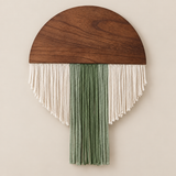 A mushroom-shaped wall hanging with a dark brown wooden cap and fringe tassels in off-white and sage green, displayed against a solid beige background.