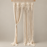 An off-white macrame curtain panel hanging from a wooden dowel, featuring intricate woven geometric patterns and long fringe tassels, set against a solid neutral beige background.